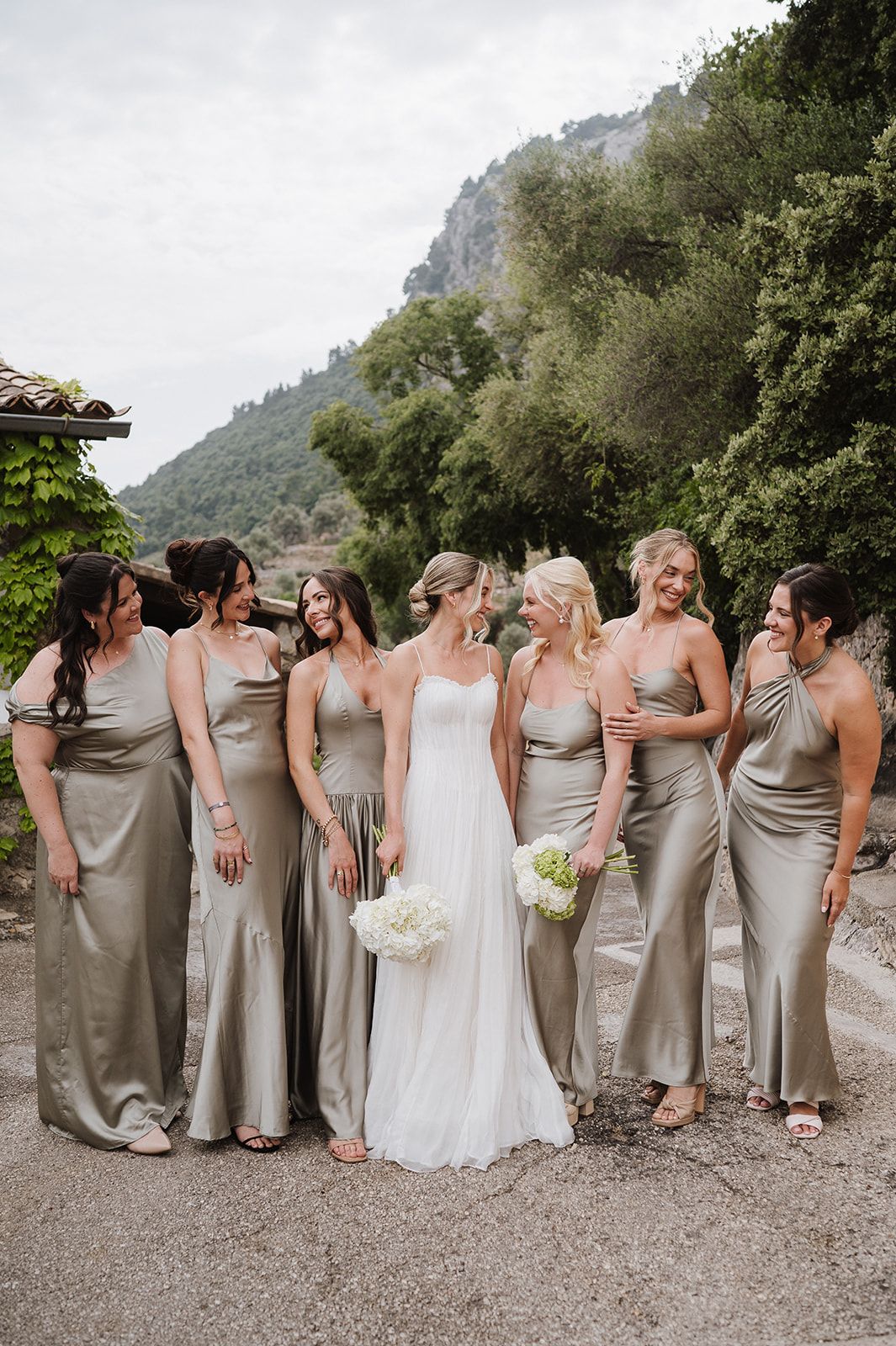 Bride holding white bouquet poses with six bridesmaids in elegant taupe satin gowns with mountain scenery behind them.