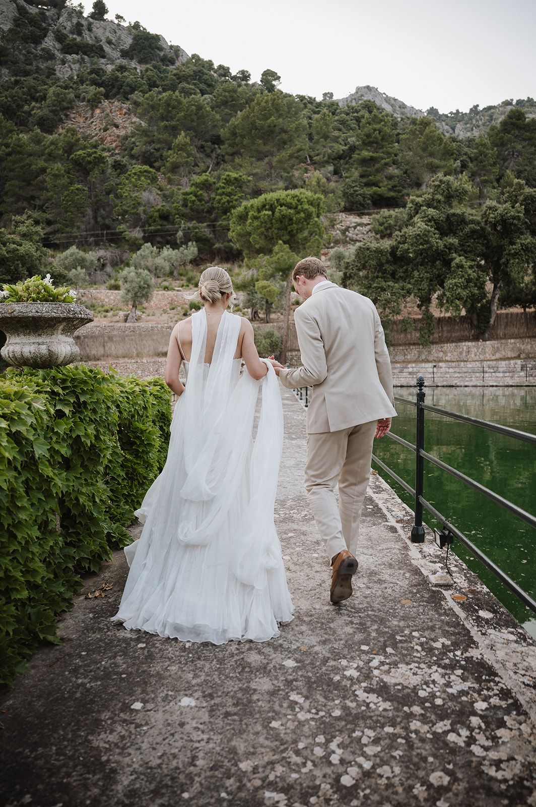 Bride in flowing white gown and groom in beige suit walk along stone path beside green reflective pond at wedding venue.