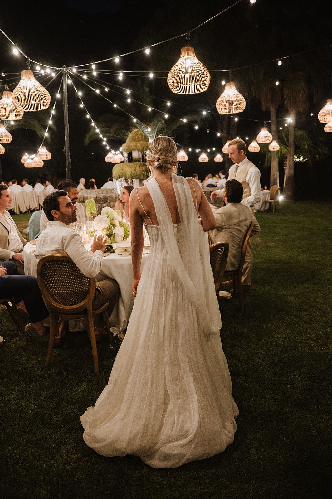 Bride in flowing white gown stands at elegant outdoor night reception under warm glowing pendant lights.