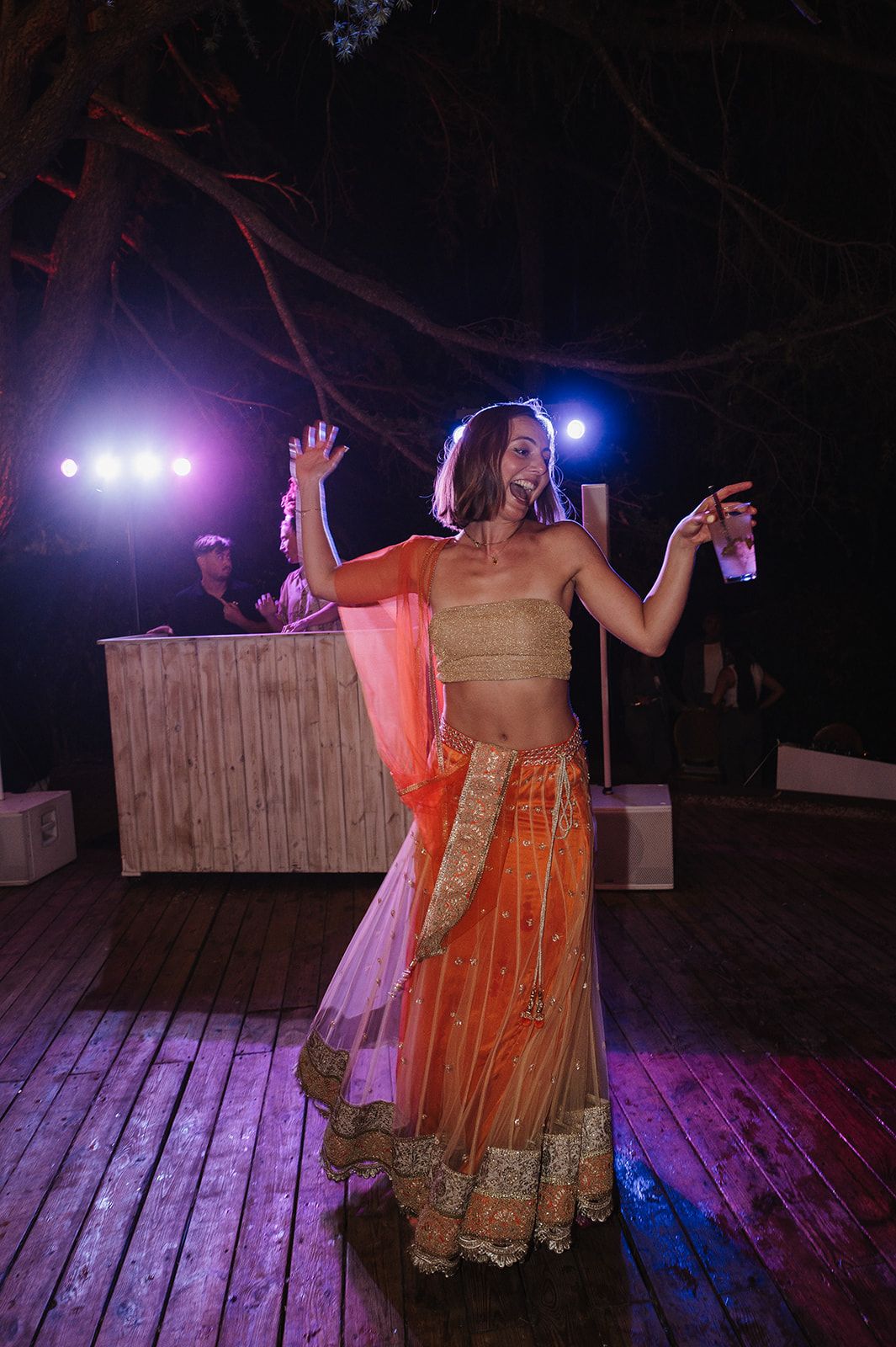 Woman in orange and gold lehenga dances joyfully on outdoor dance floor at night during Indian wedding.