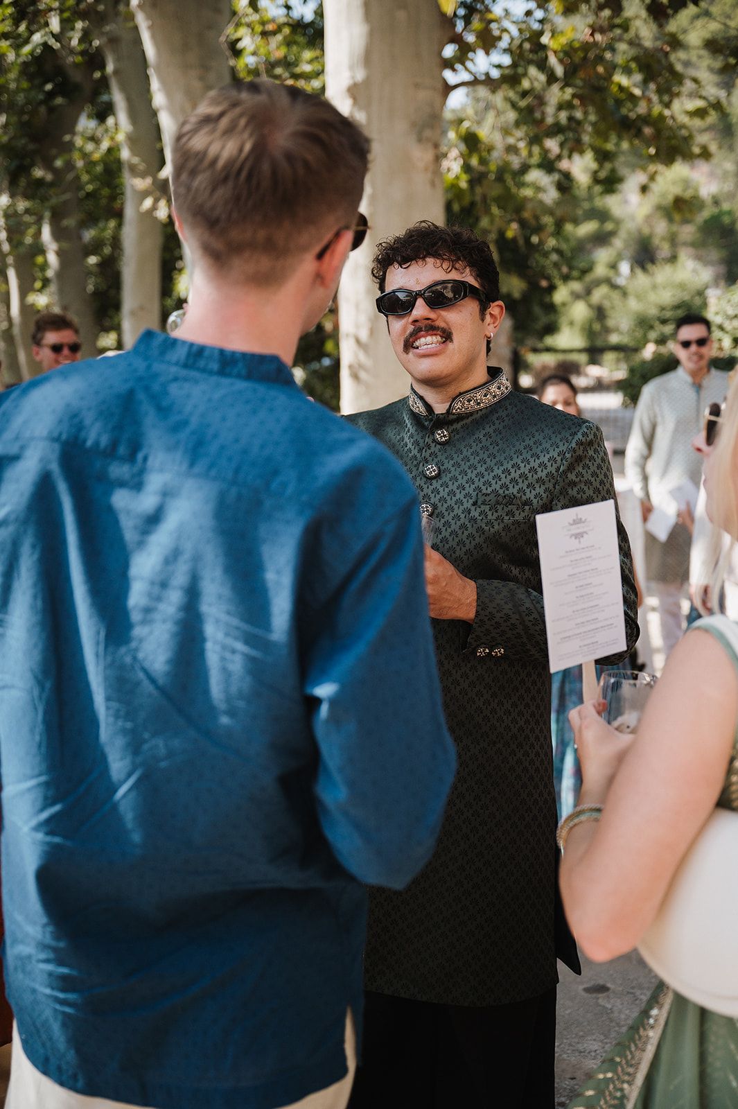 Man in dark green sherwani with sunglasses holding ceremony program at outdoor wedding.