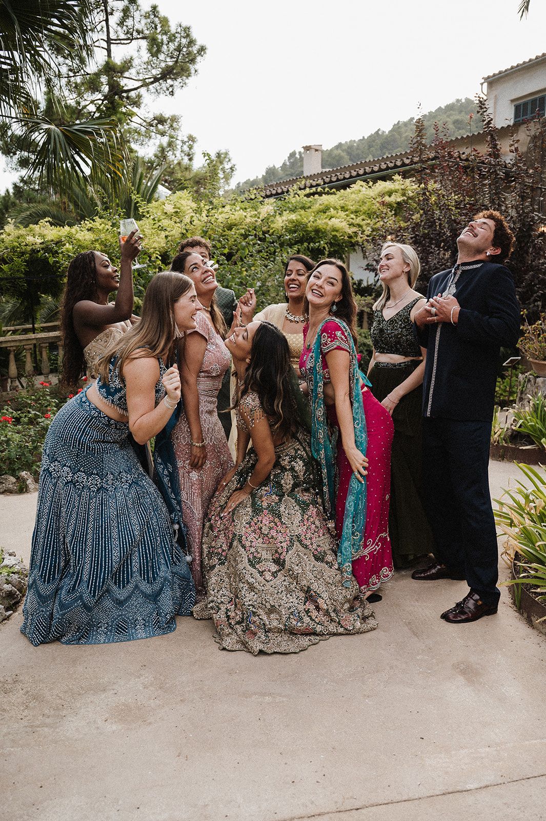 Wedding guests in vibrant Indian outfits pose and laugh together in a lush garden setting.