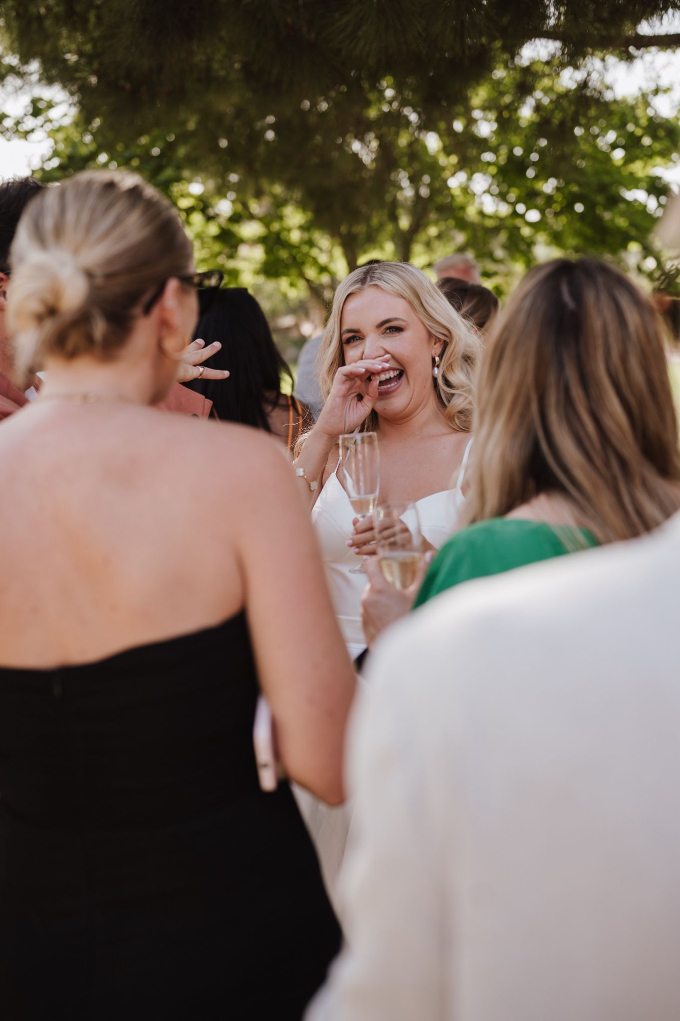 Laughing bride holding champagne glass mingling with guests at outdoor wedding reception.