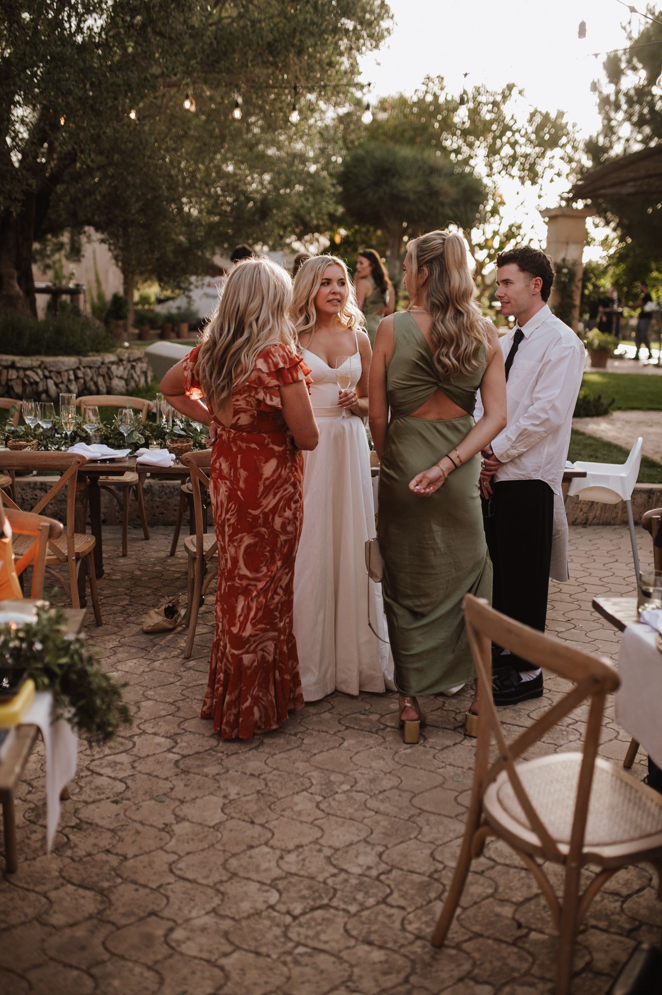 Bride in white gown chats with guests in red floral and green dresses at outdoor wedding reception at dusk.