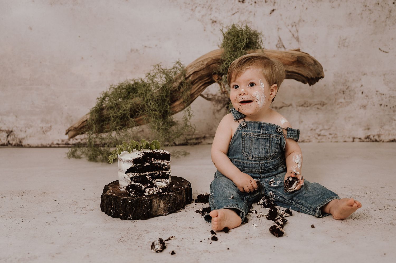 Baby in denim overalls smiling beside a smashed chocolate cake on a white studio floor.