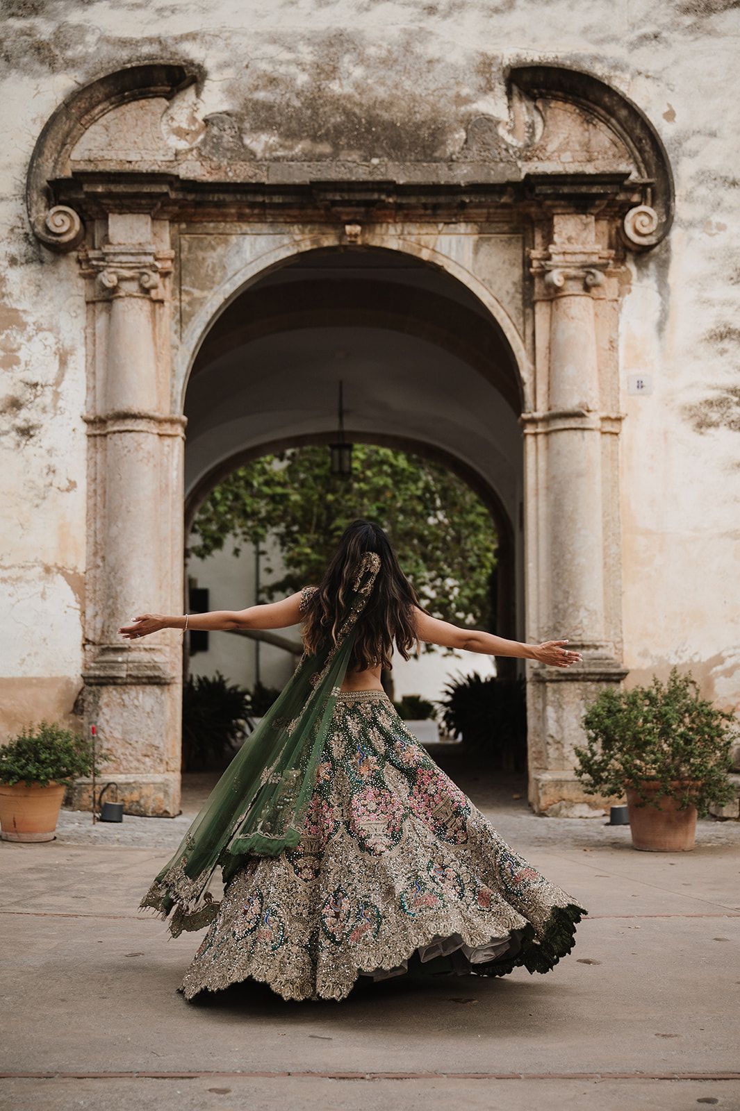 Woman twirling in ornate green floral lehenga before a grand stone archway.