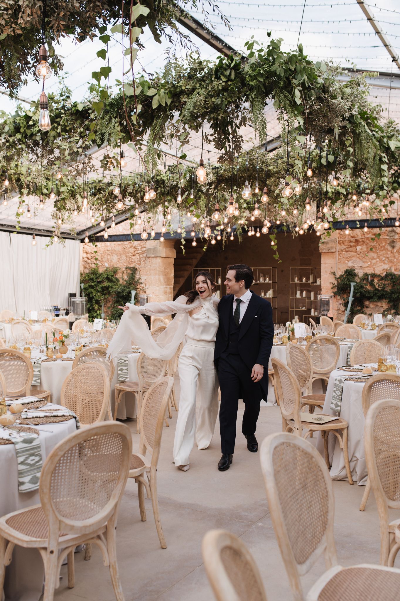 Bride and groom walk through elegant wedding reception with rattan chairs and lush floral ceiling installation.