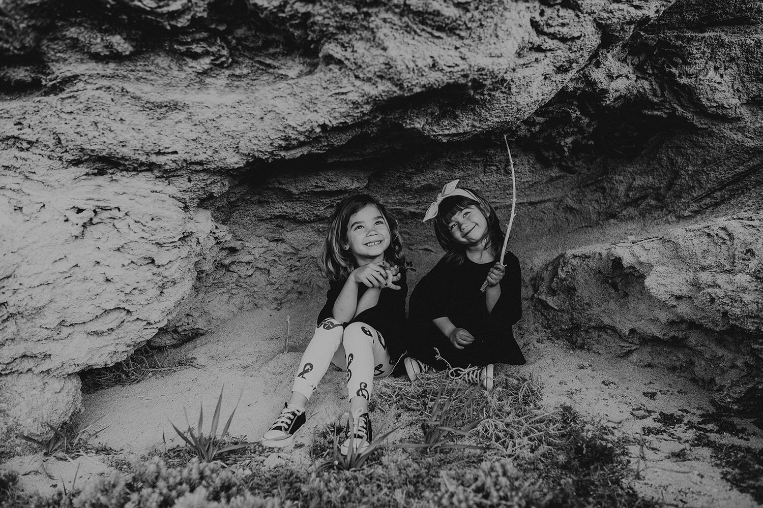 Two young girls smile together nestled in a rocky cave in this charming black and white photo.