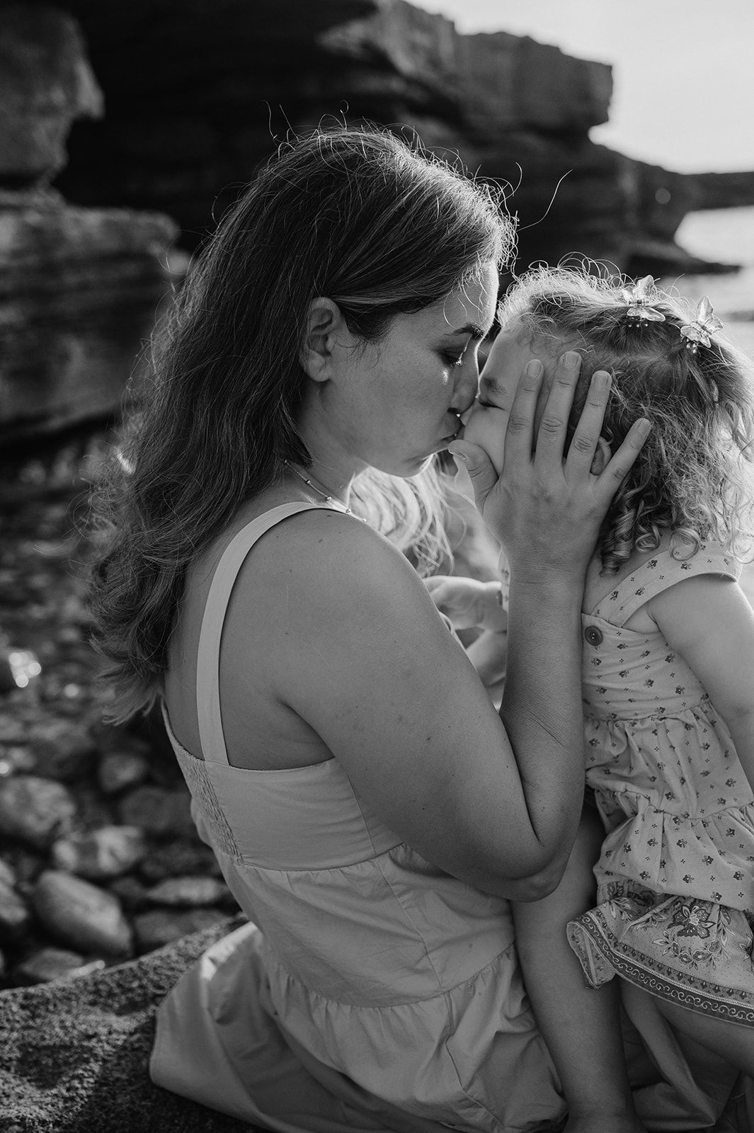 Black and white image of a mother tenderly kissing her young daughter outdoors.