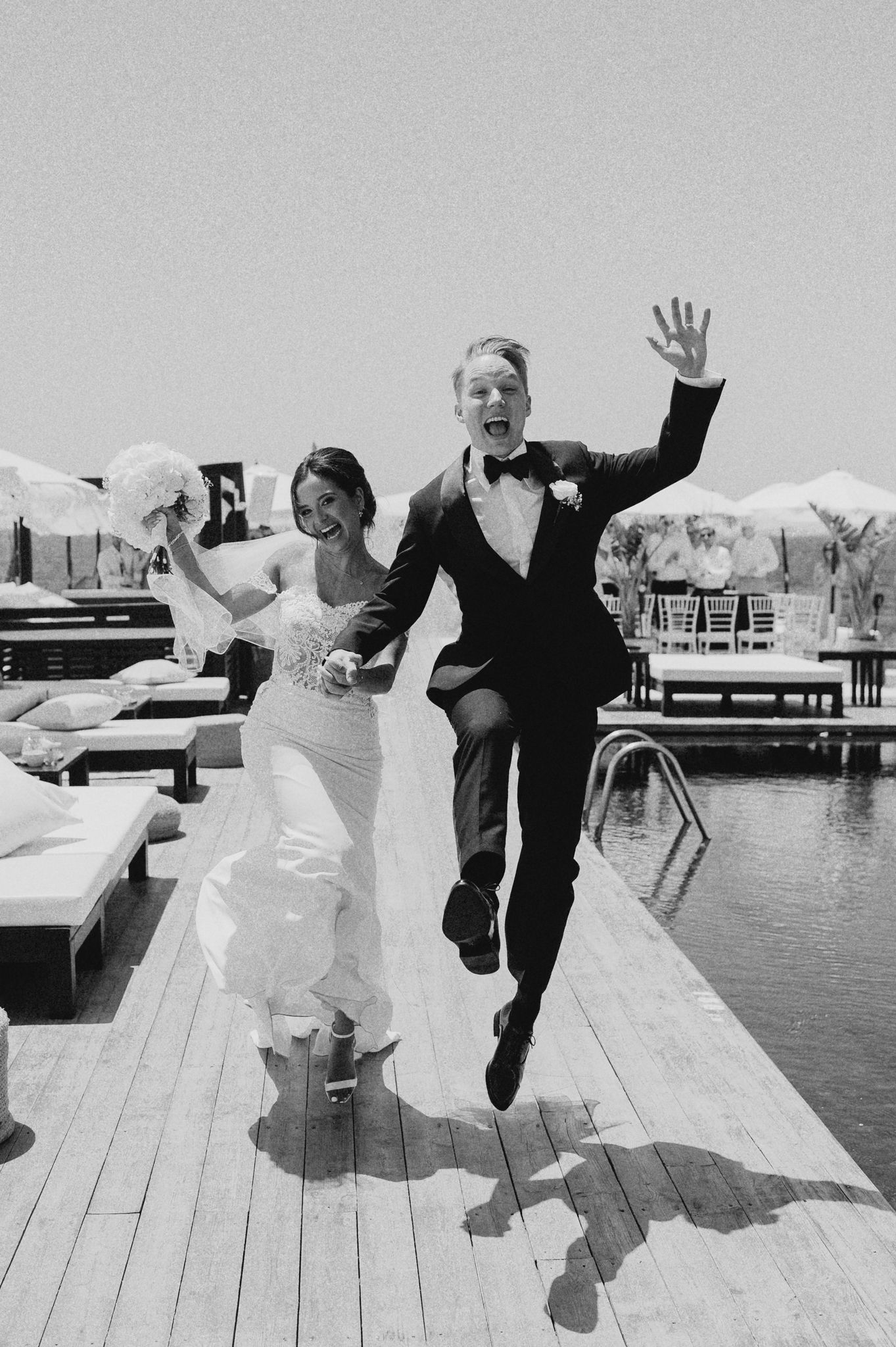 Black and white photo of joyful newlyweds jumping and celebrating on a poolside deck after their wedding ceremony.