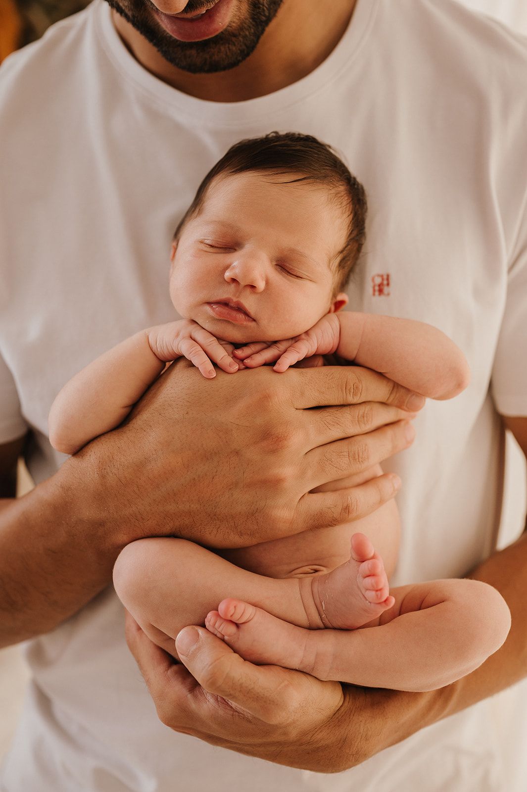 Father gently cradling sleeping newborn baby in both hands, wearing white t-shirt.