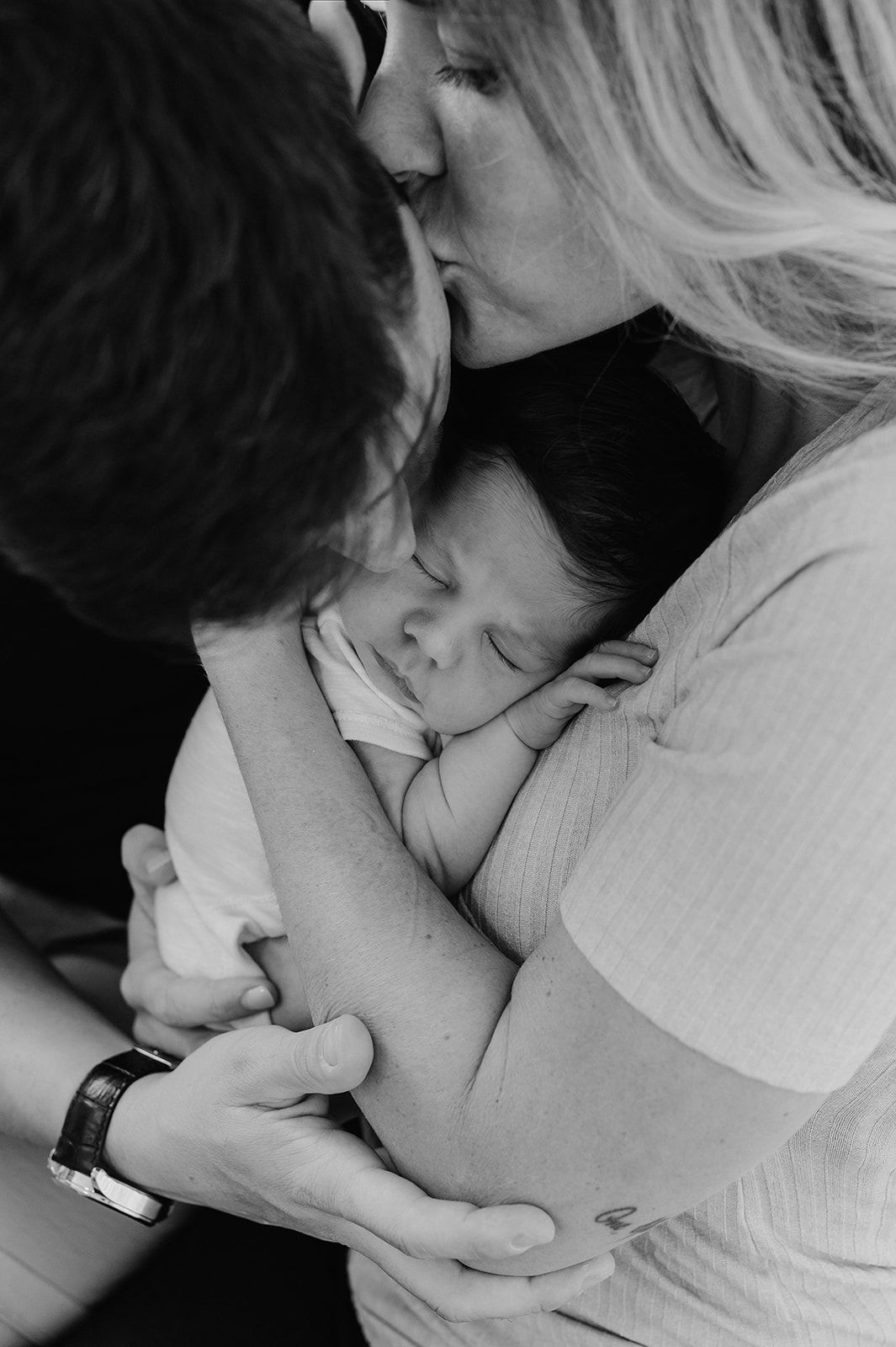 Black and white photo of new parents kissing while cradling their sleeping newborn baby together.