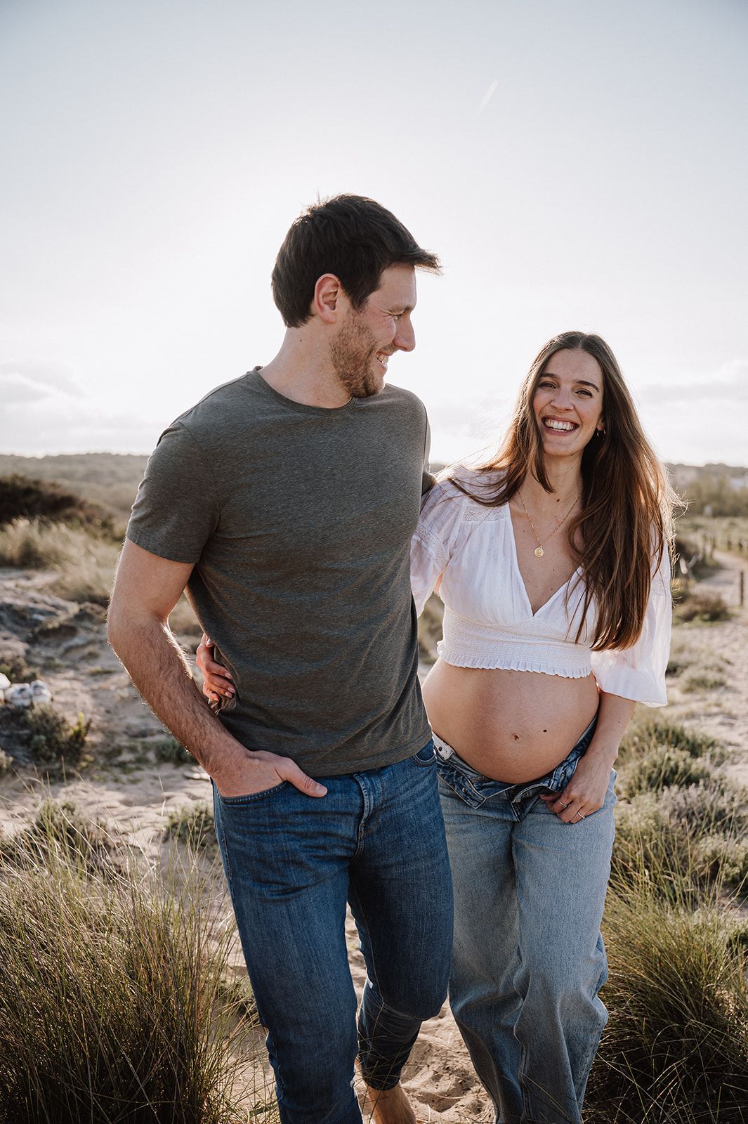 Happy pregnant couple laughing together outdoors at sunset, woman showing baby bump in white crop top and jeans.