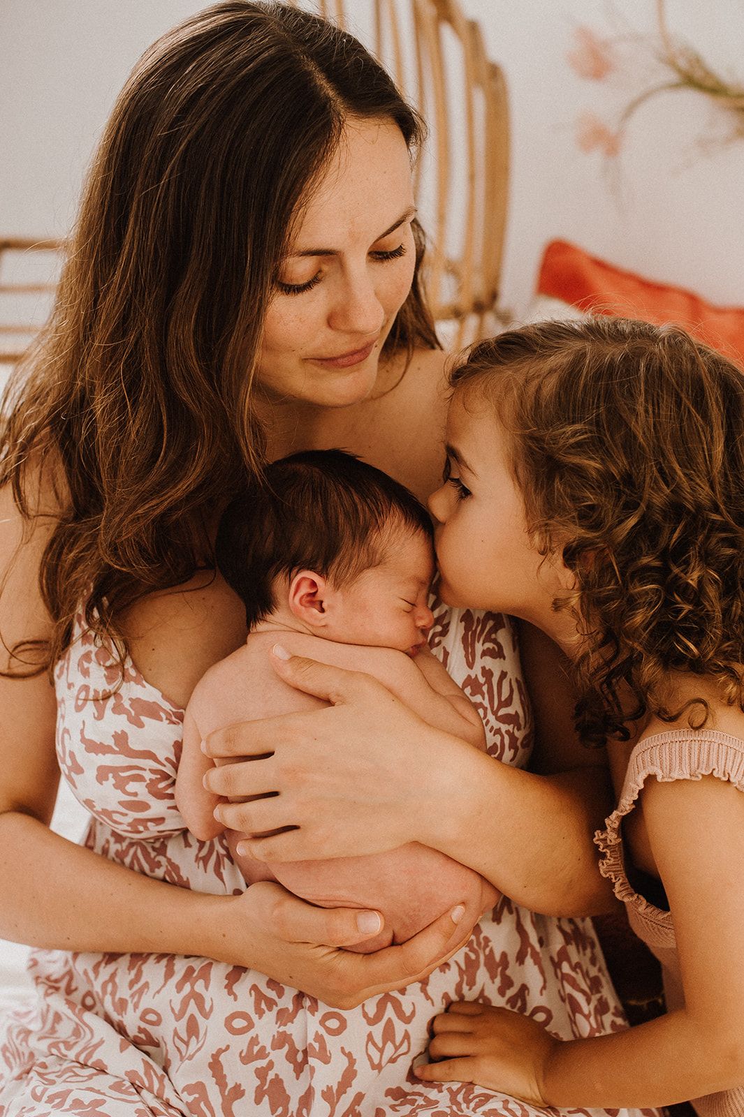 A mother tenderly holds her newborn while her young daughter gives the baby a gentle kiss.
