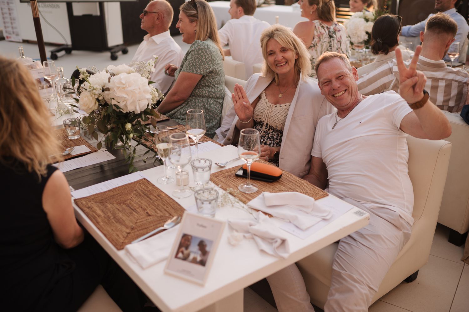 Wedding guests in white attire smile and celebrate at outdoor reception dinner table.