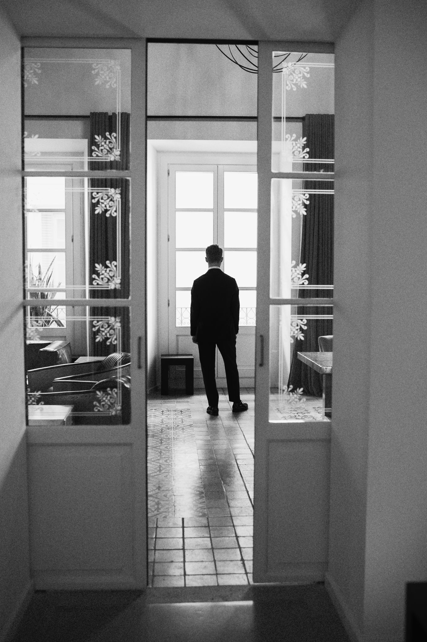 Groom in black suit stands alone by window in black and white photo.