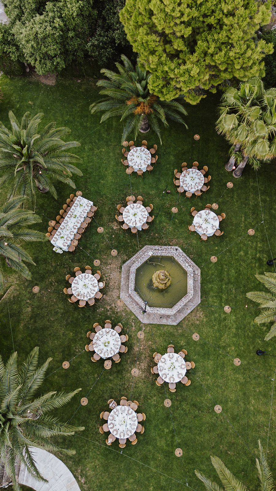 Aerial view of outdoor wedding reception with round tables surrounding an octagonal fountain in a lush garden.