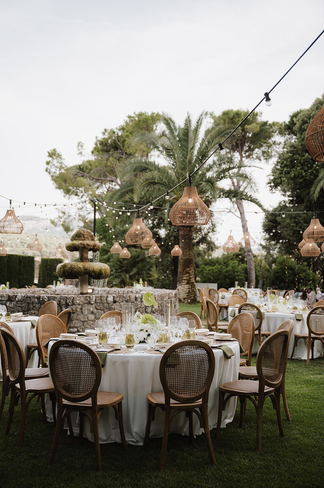 Elegant outdoor wedding reception with rattan pendant lights, cane chairs, and white linen tables on a lawn.