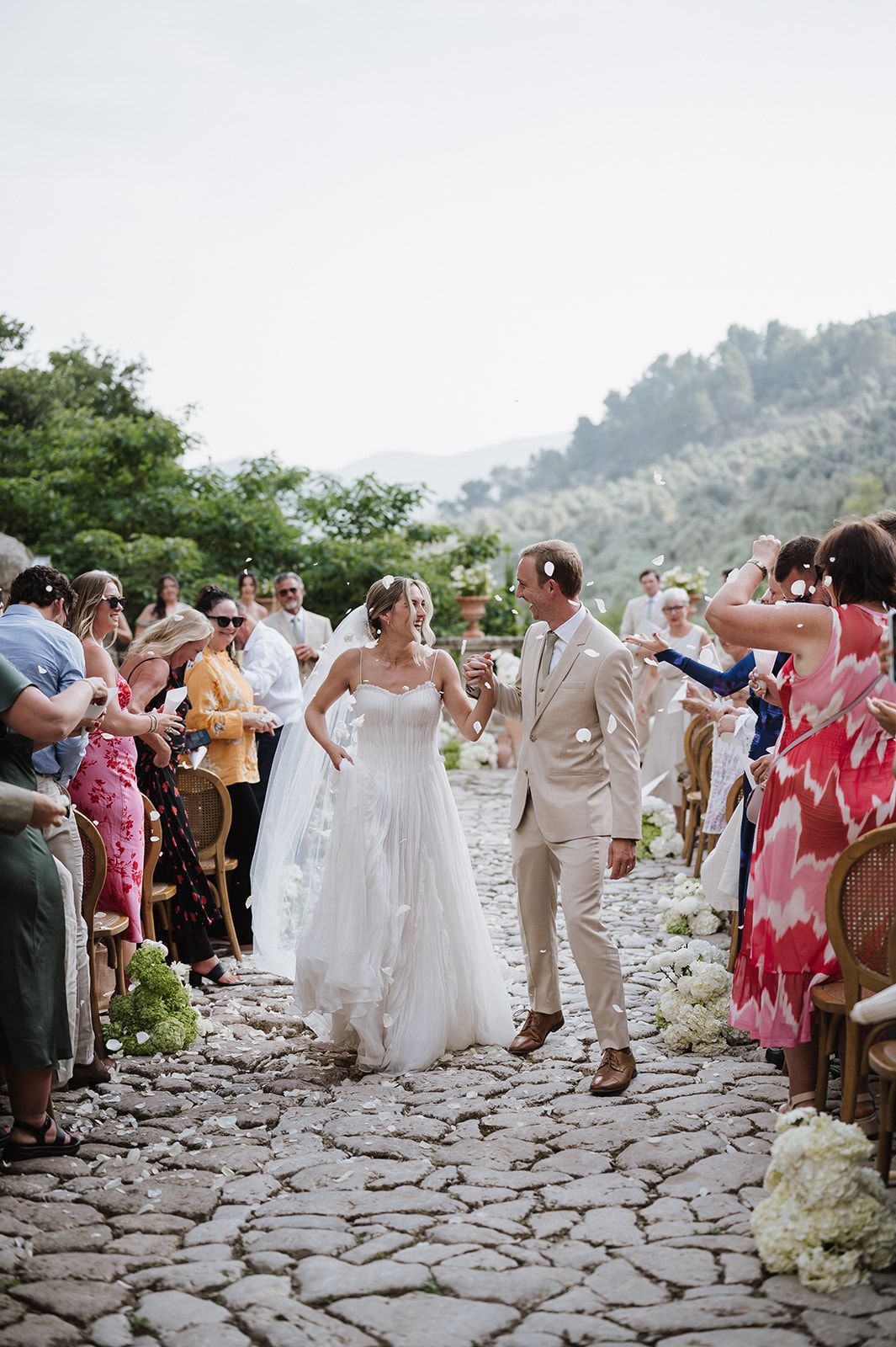 Newlyweds walk joyfully down cobblestone aisle as guests celebrate at outdoor Mediterranean wedding.