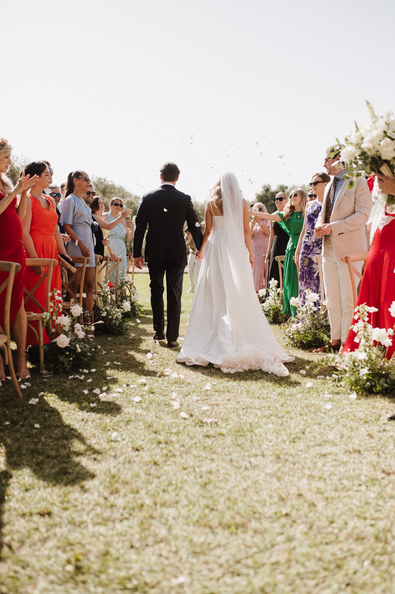 Newlyweds walk back down aisle as guests toss confetti petals at sunny outdoor garden wedding ceremony.