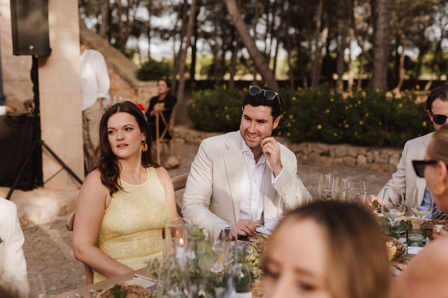 Wedding guests in colorful attire enjoy conversation and drinks at outdoor garden reception dinner table.