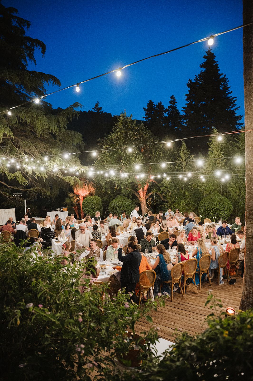 Outdoor wedding reception at dusk with string lights over long dining tables surrounded by lush greenery.