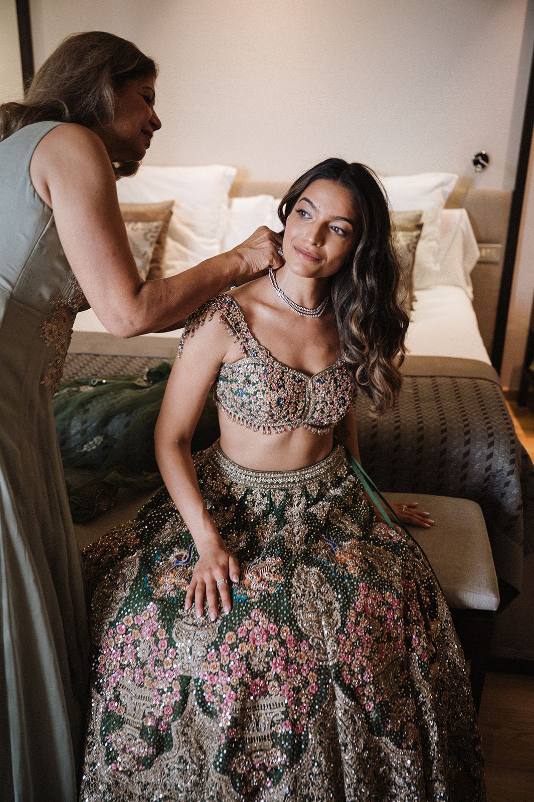 Bride in ornate green floral lehenga being helped with necklace while seated in bridal suite.