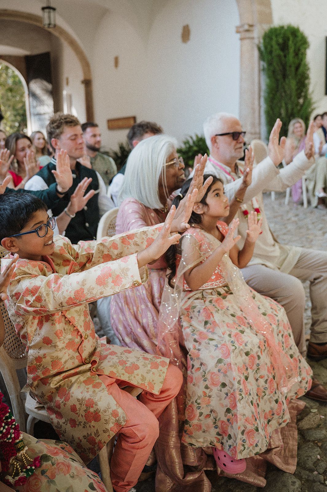 Wedding guests in floral Indian outfits clapping joyfully during outdoor Hindu wedding ceremony.
