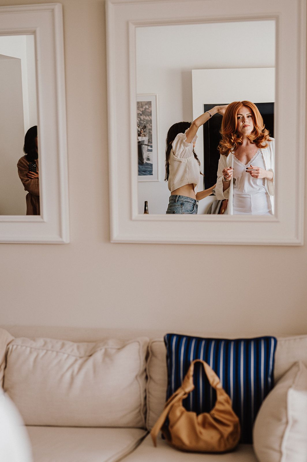Red-haired bride getting ready reflected in white-framed mirrors above couch with striped pillow.