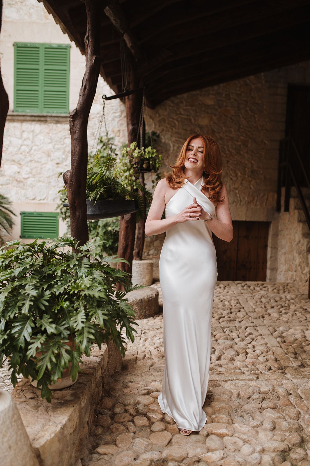 Elegant bride in white halter neck satin gown standing in a cobblestone Mediterranean courtyard.