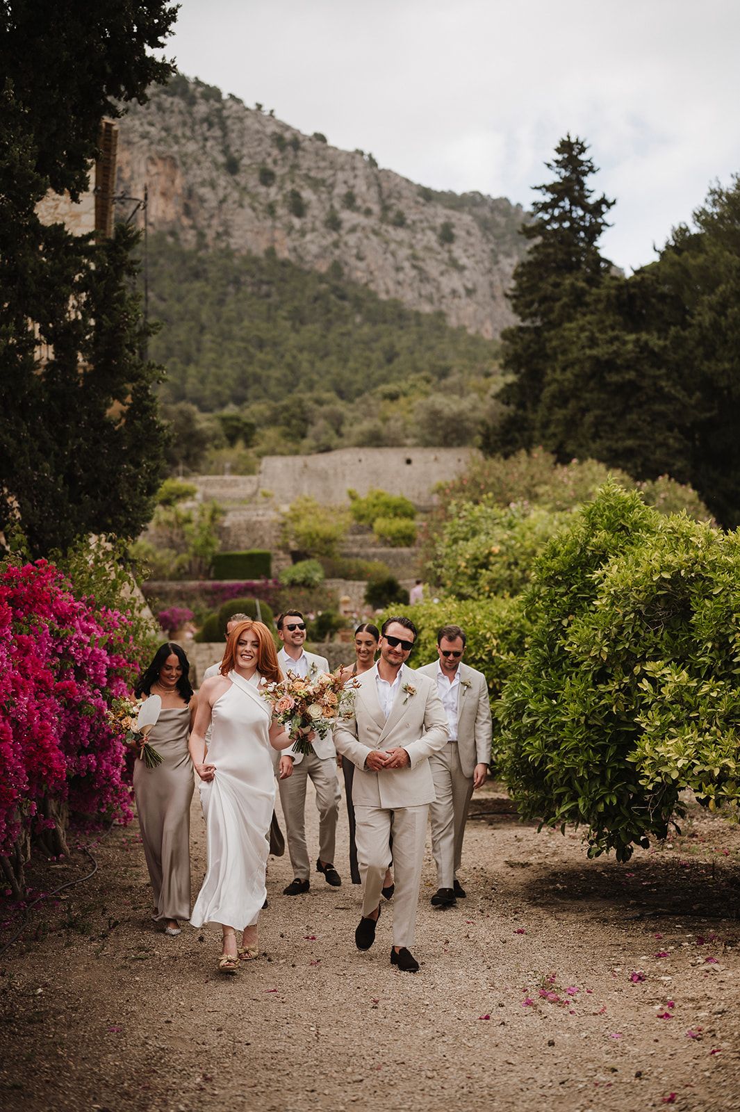 Bride and groom lead wedding party through lush garden with dramatic mountain backdrop.