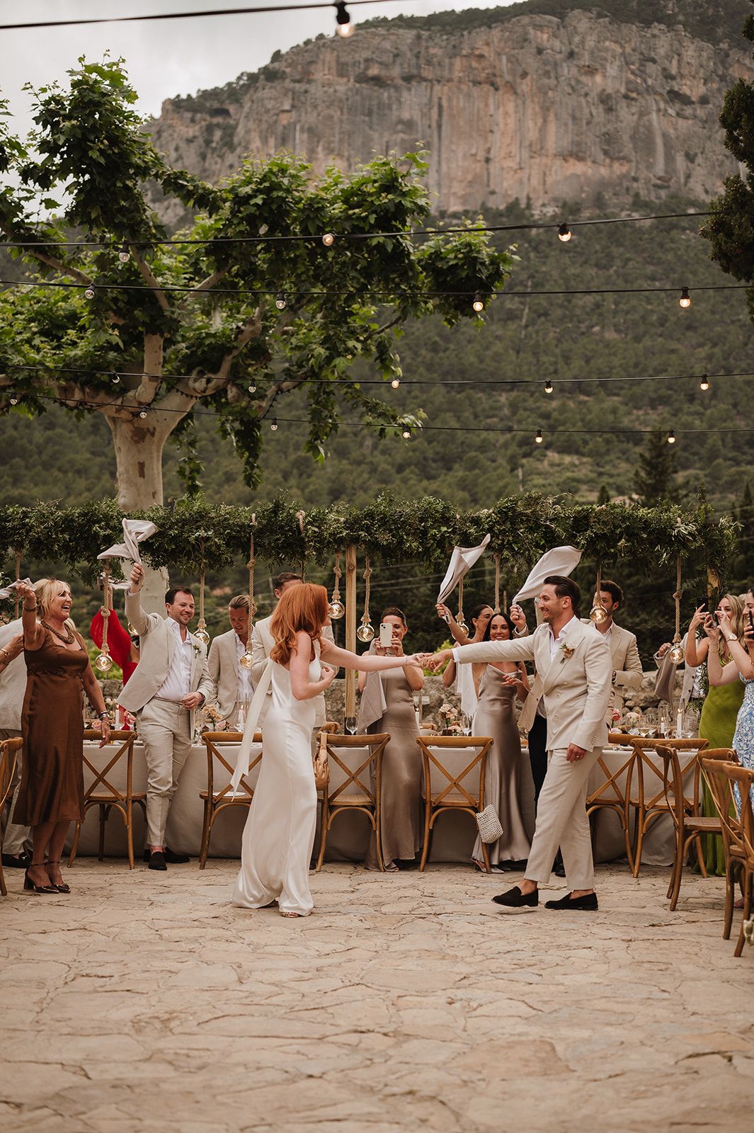 Bride and groom dance joyfully outdoors at a rustic Mediterranean wedding reception with string lights and mountain backdrop.