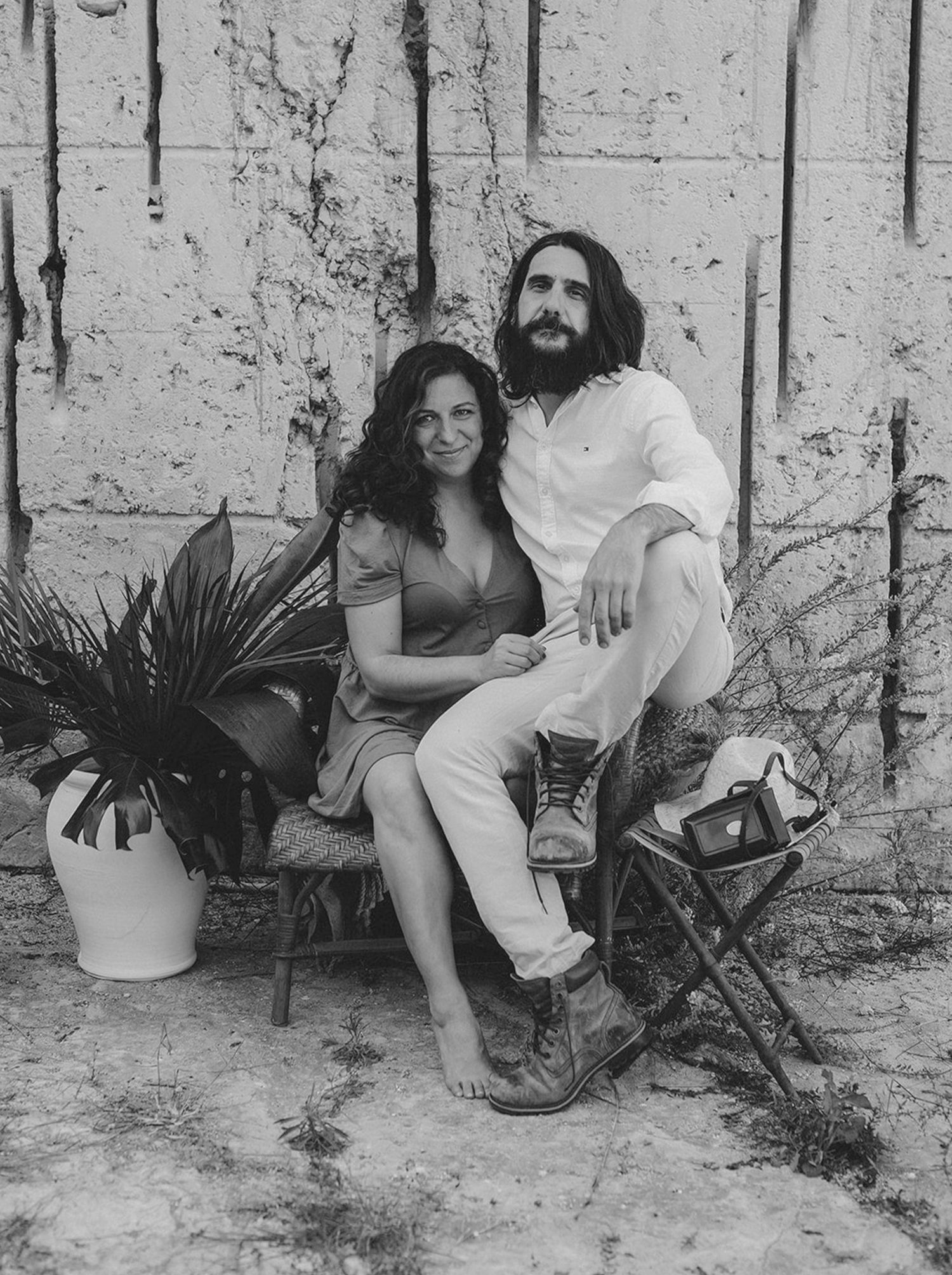 Black and white portrait of couple sitting together against rustic stone wall with plants.