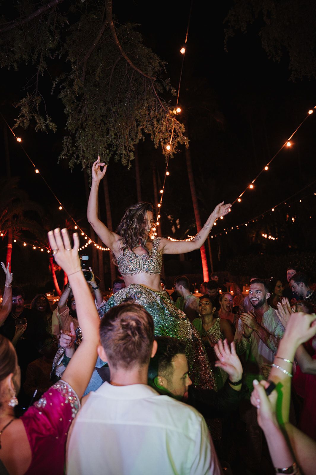 Bride in embellished lehenga lifted by guests during lively outdoor wedding celebration at night.