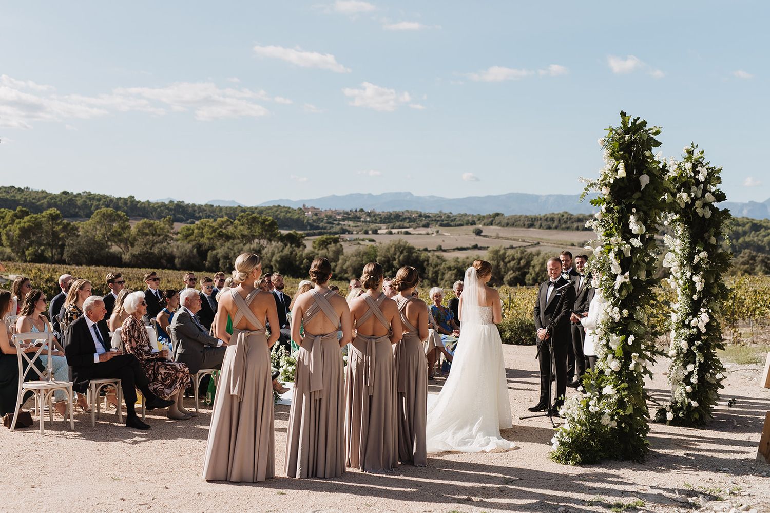 Outdoor wedding ceremony with bridesmaids in taupe gowns, floral arch, and scenic vineyard backdrop.