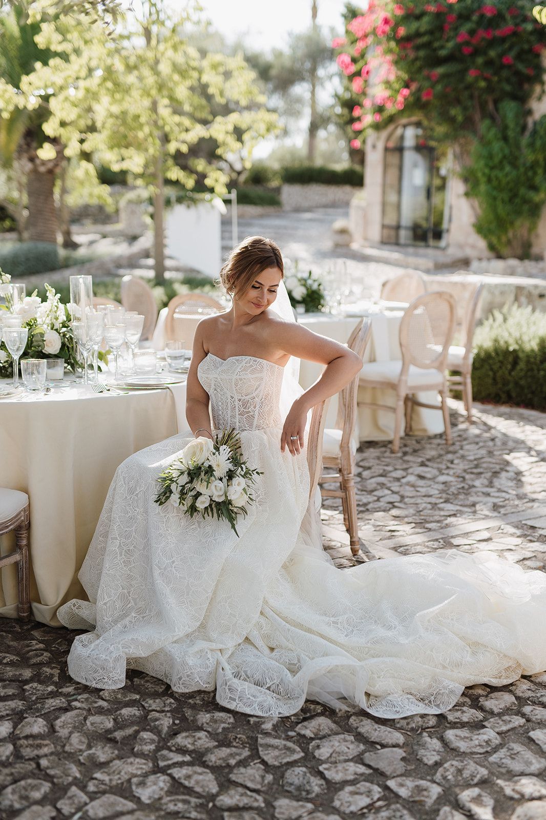 Bride in strapless lace mermaid gown sits gracefully beside reception tables holding a white bouquet.