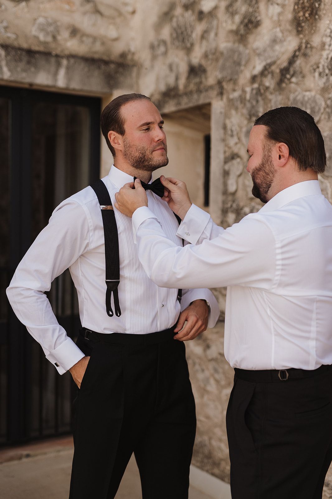 Groomsman adjusting bow tie of groom wearing suspenders before wedding ceremony outdoors.