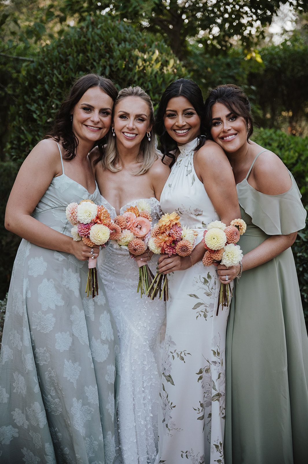 Bride with three bridesmaids in sage green dresses holding coral and white floral bouquets outdoors.