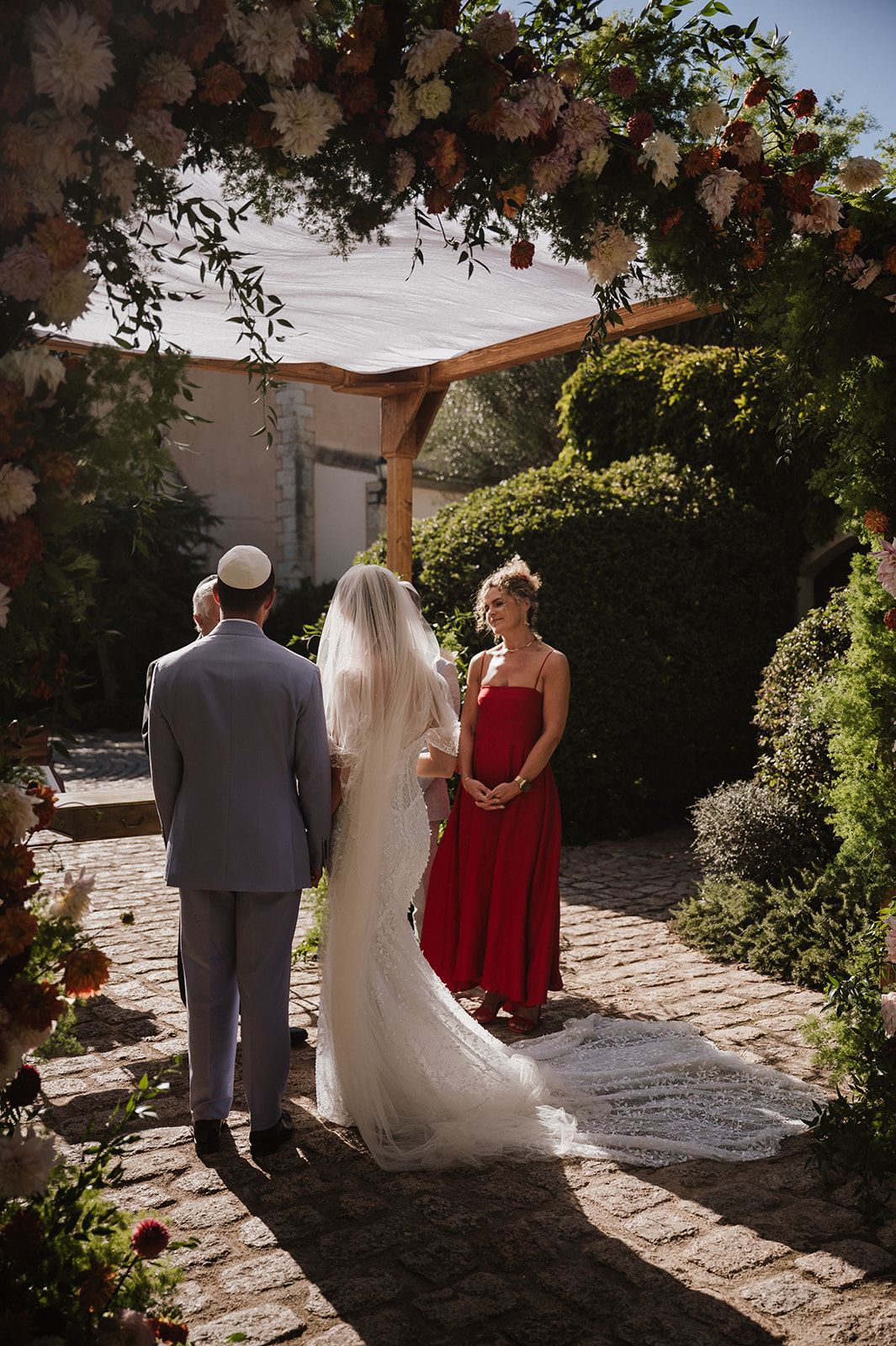 Bride and groom stand before officiant under floral arch during outdoor Jewish wedding ceremony in garden.
