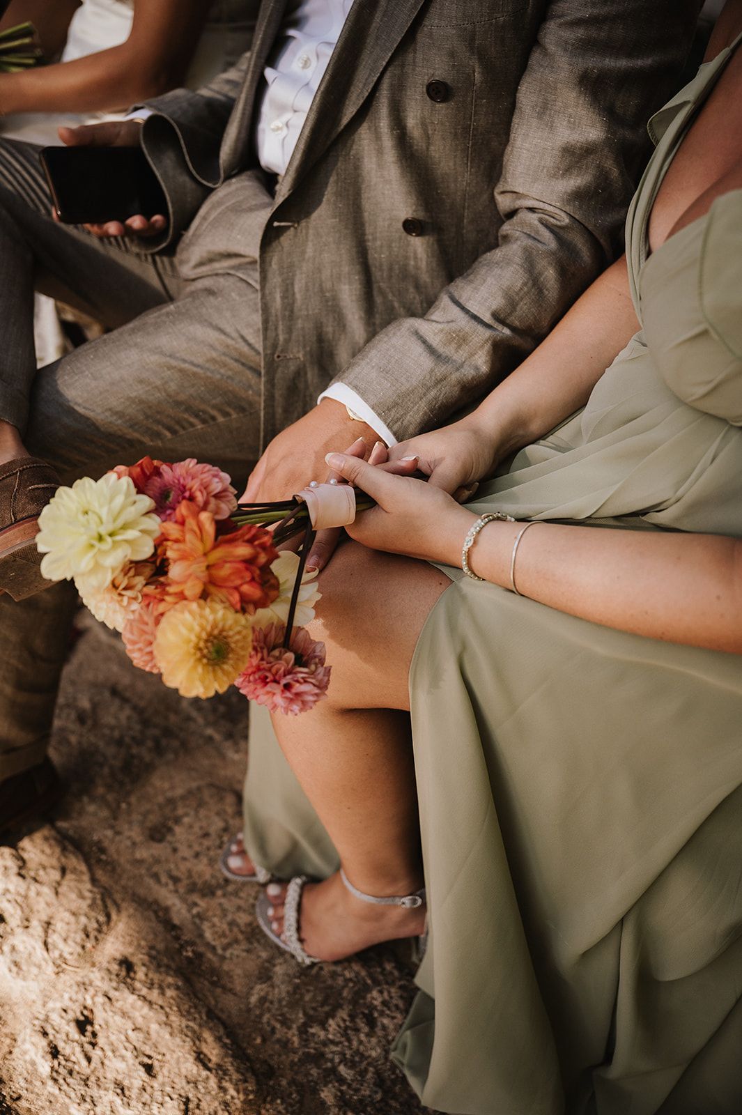 Wedding guest in sage green dress holding a colorful bouquet of dahlias and zinnias while seated at the ceremony.