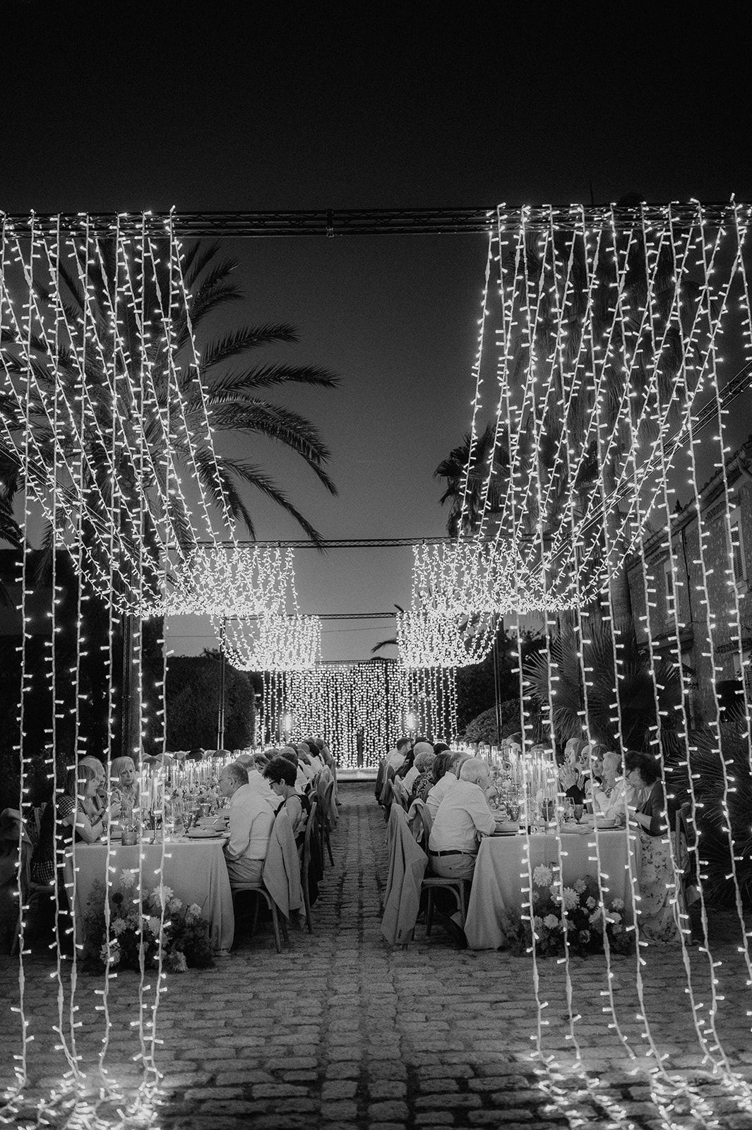 Black and white aerial view of outdoor wedding reception illuminated by cascading fairy light curtains.