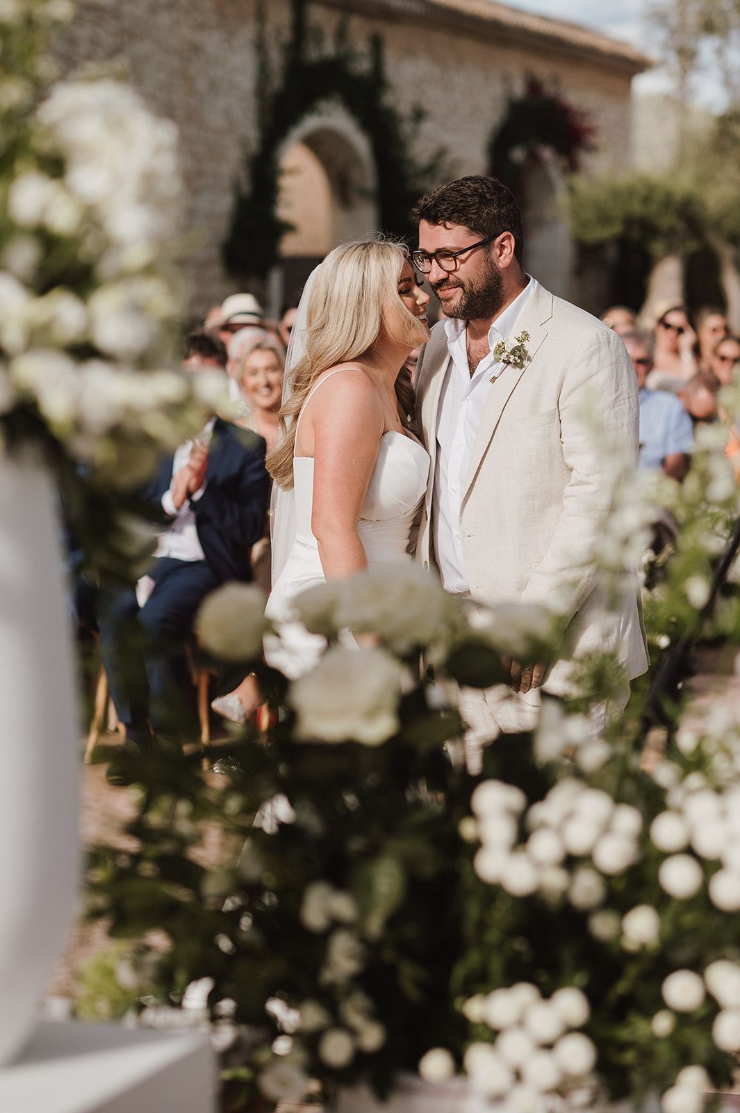 Bride and groom share a joyful moment at the altar surrounded by lush white floral arrangements.