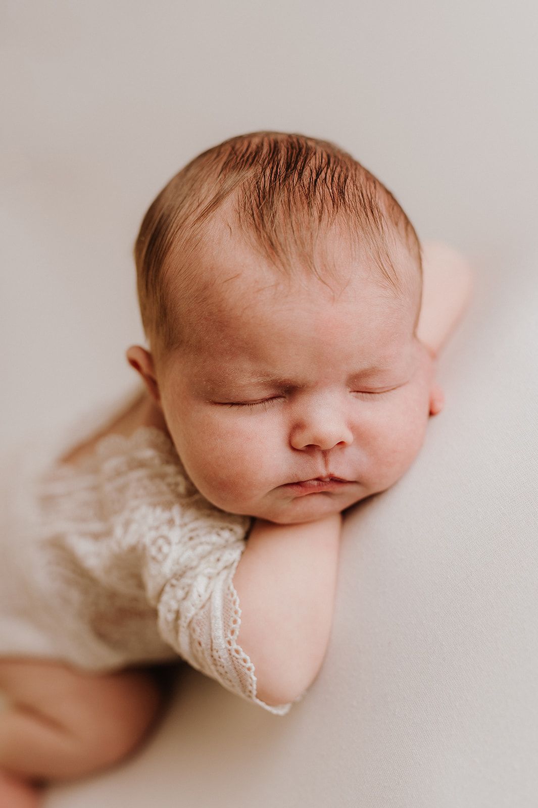 Sleeping newborn baby in lace romper resting head on hands against soft cream background.