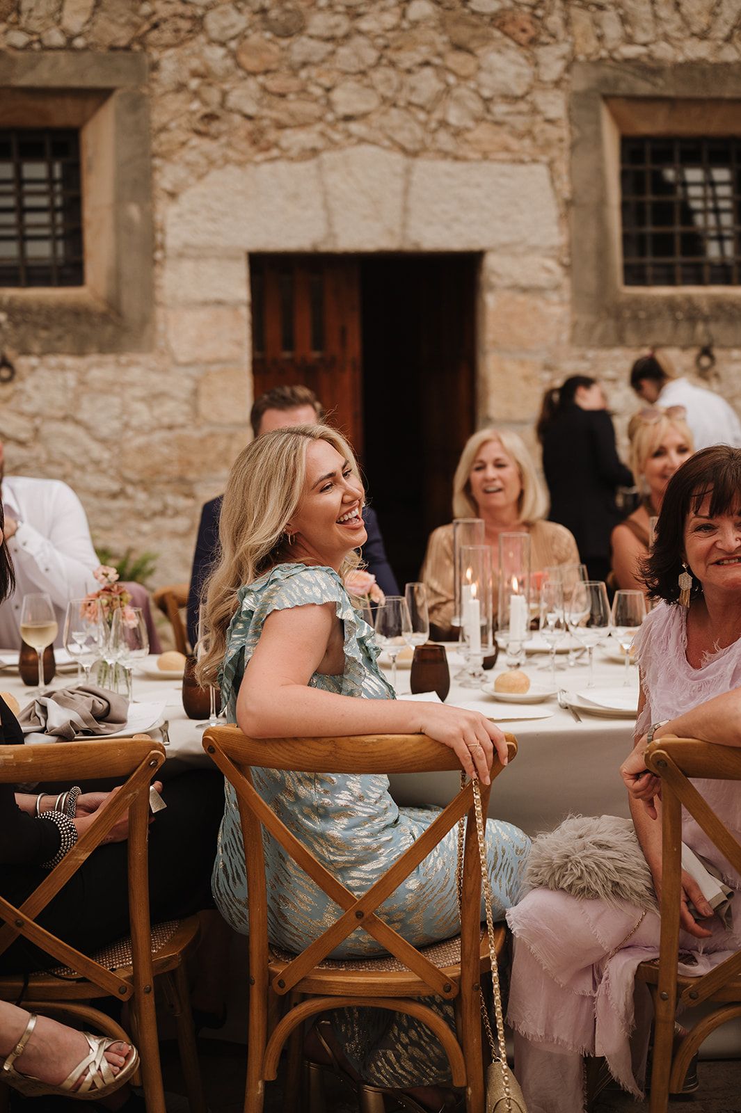 Blonde wedding guest in floral dress smiles at outdoor stone venue reception dinner table.