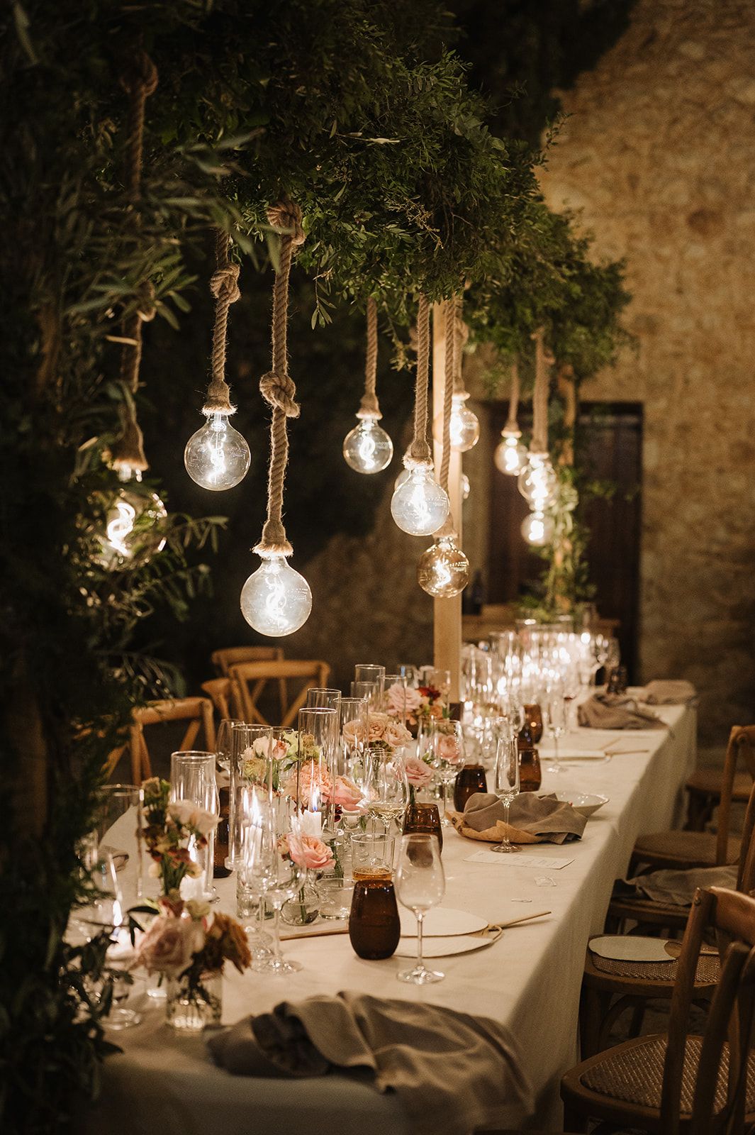 Elegant wedding reception table with hanging Edison bulbs, pink florals, candles, and lush greenery.