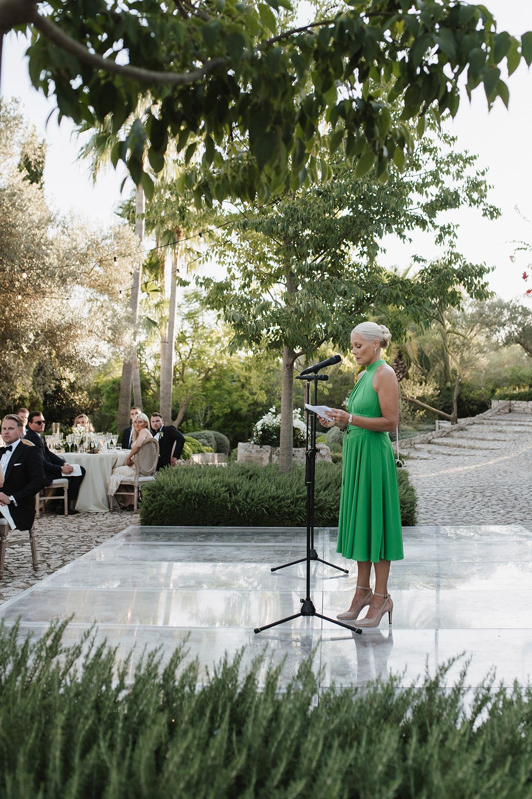 Woman in green dress delivers a speech at a microphone on a dance floor during an outdoor wedding reception.