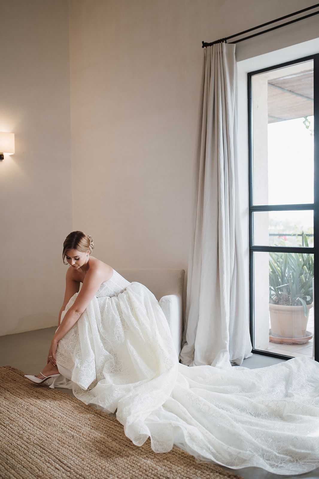 Bride in elegant white lace wedding gown putting on heels in bright room.