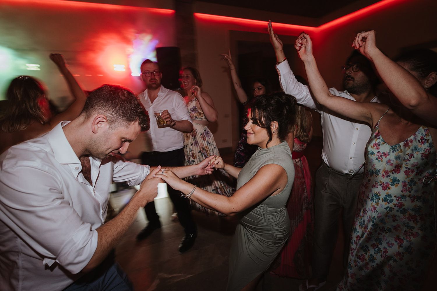 Wedding guests dance energetically on a colorful lit dance floor at a reception.