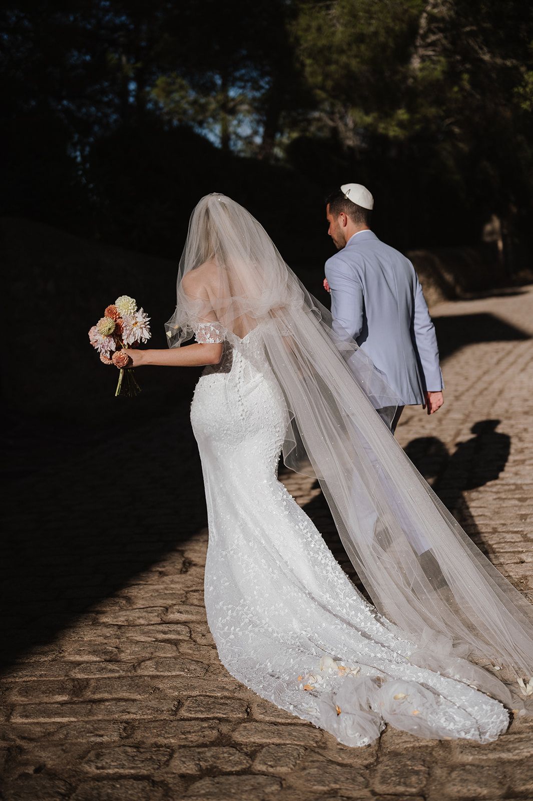 Bride in lace mermaid gown with long veil holding bouquet walks with groom in gray suit on cobblestone path.