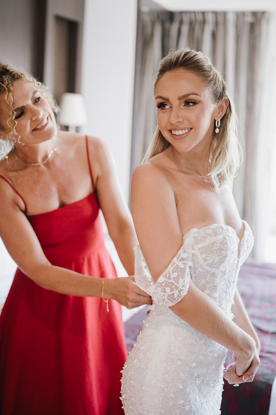 Bride in strapless lace wedding gown being helped by woman in red dress.